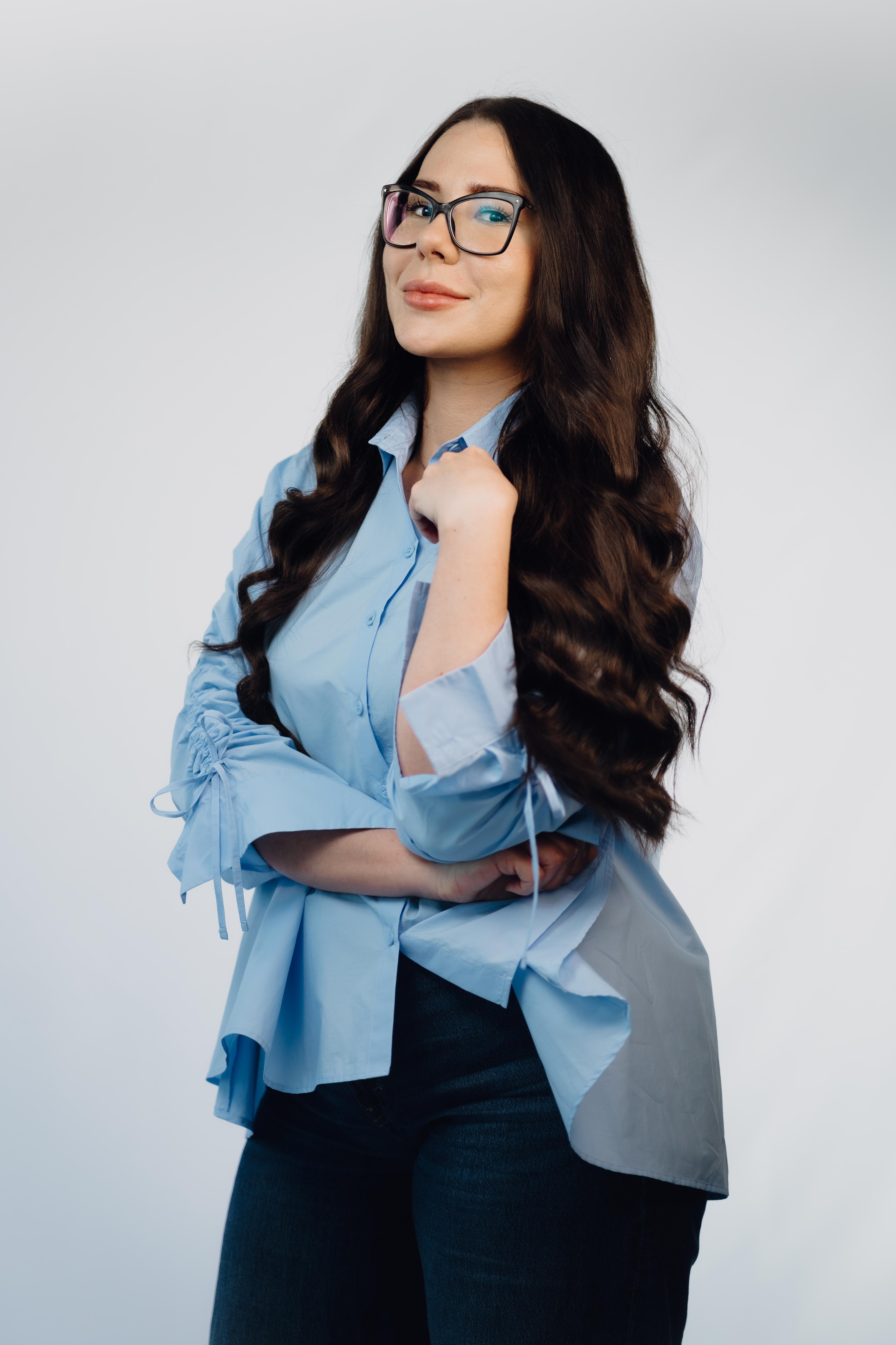 Professionelles Porträtfoto einer Frau mit langen dunklen gewellten Haaren und Brille, in einer hellblauen Bluse und dunkler Jeans, mit verschränkten Armen und leichtem Lächeln in die Kamera blickend.