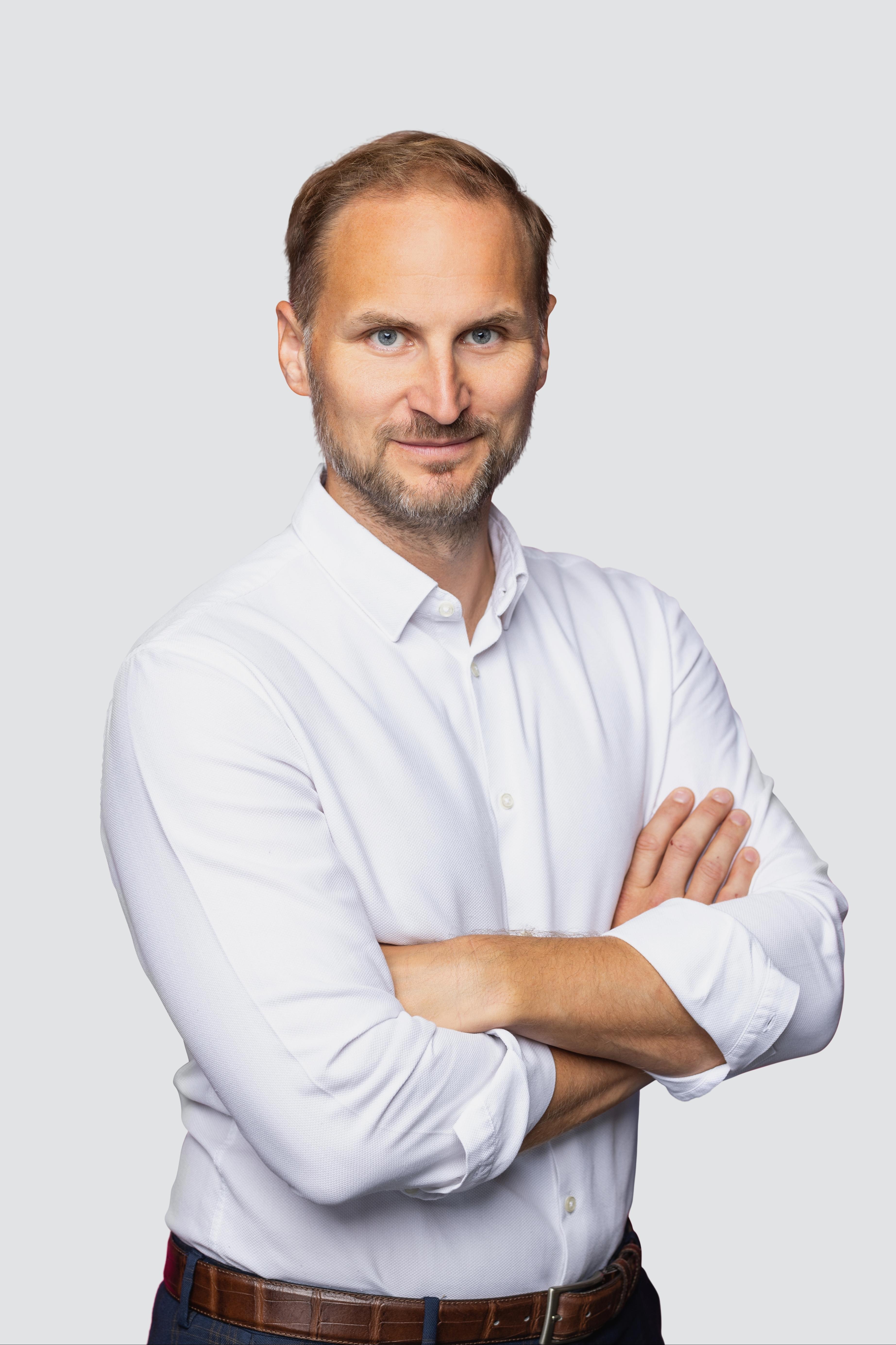 Professionelles Porträtfoto eines Mannes mit kurzen rotbraunen Haaren und leichtem Bart, in einem weißen Hemd mit hochgekrempelten Ärmeln, mit verschränkten Armen in die Kamera blickend.