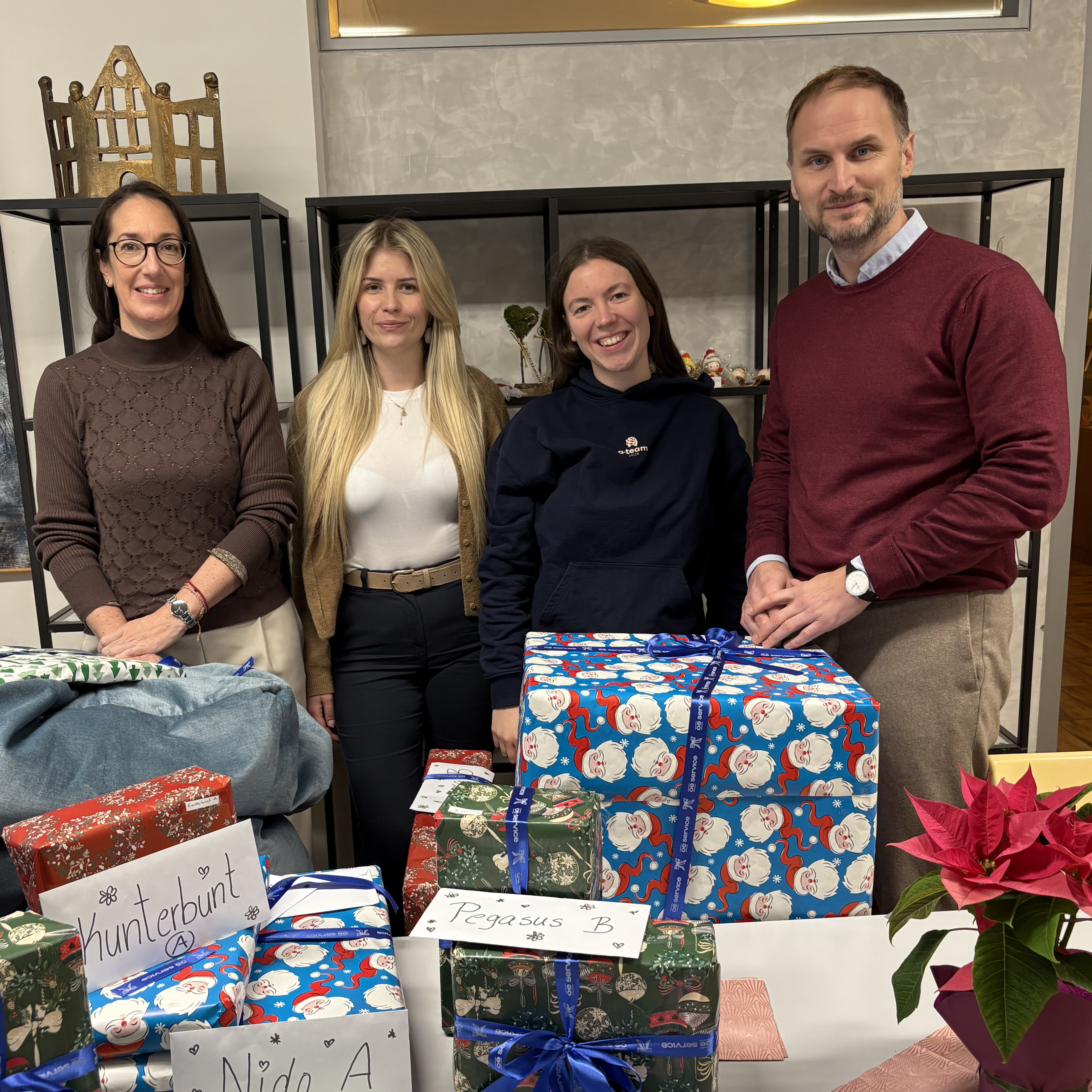 Vier Kollegen stehen hinter einem Tisch voller verpackter, mit Namen beschrifteter Weihnachtsgeschenke und lächeln in die Kamera. Das Büro hat Glasvitrinen und in der Ecke steht eine Weihnachtssternpflanze.